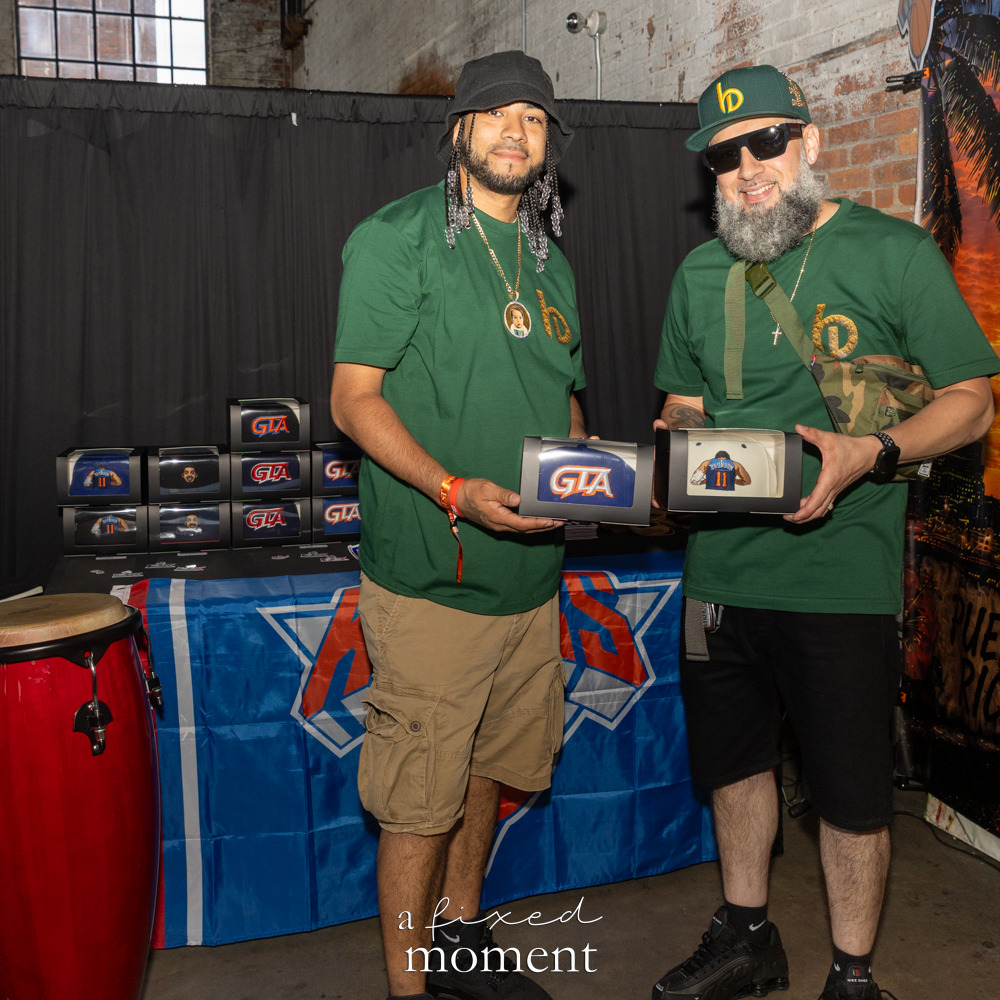 Two vendors in matching green shirts pose behind a table with boxed custom embroidery pieces, display items and a red conga drum.