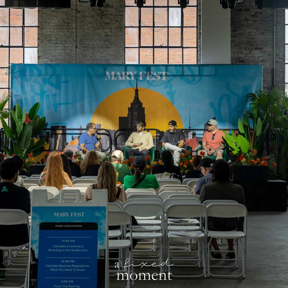 Wide view of a MARY Fest panel discussion with speakers seated onstage in front of a blue branded backdrop and an audience in white folding chairs.
