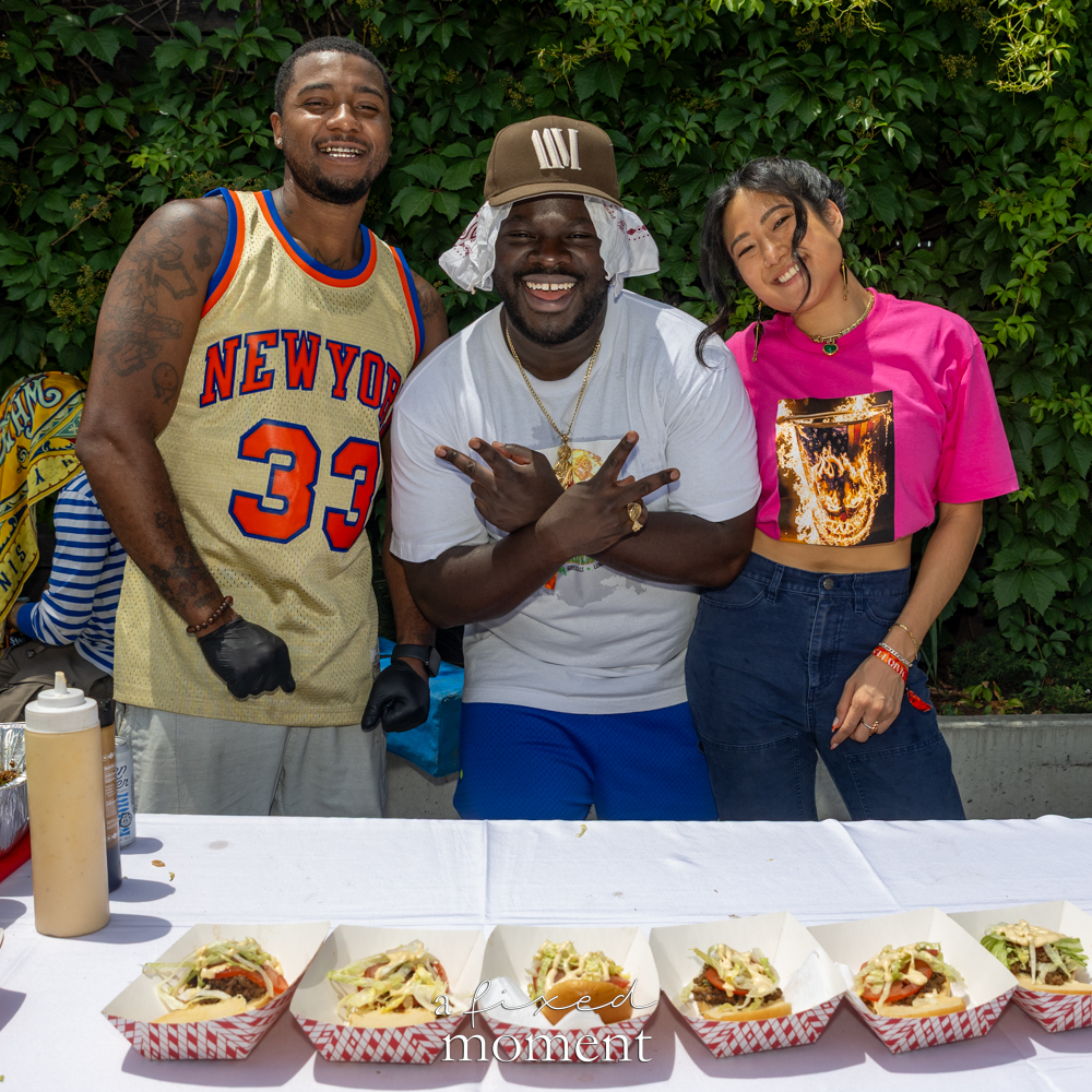 Three Shmackwich vendors pose behind a table lined with chopped cheese sandwiches in red-check paper trays.