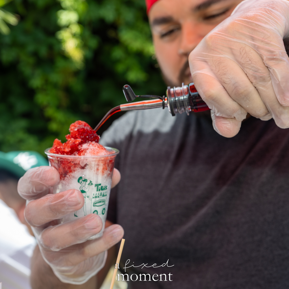 Gloved vendor pours red syrup from a bottle onto shaved ice in a small clear Titi’s cup.