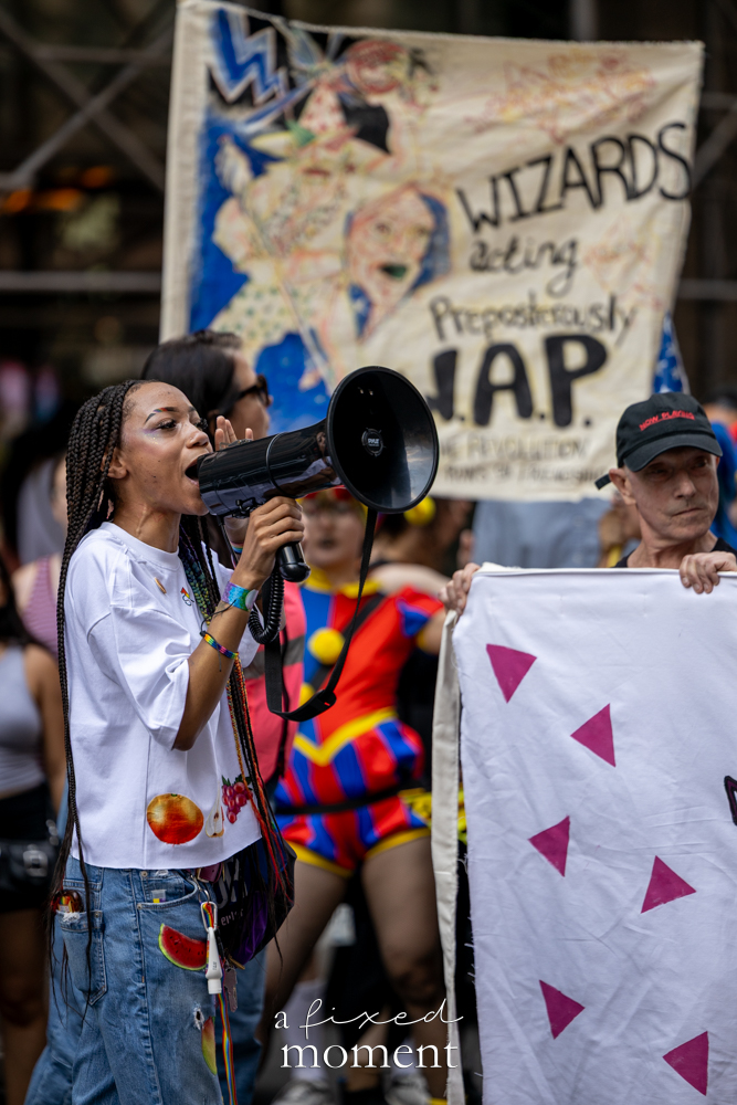Megaphone speaker at the Queer Liberation March with a Wizards Acting Preposterously banner behind them