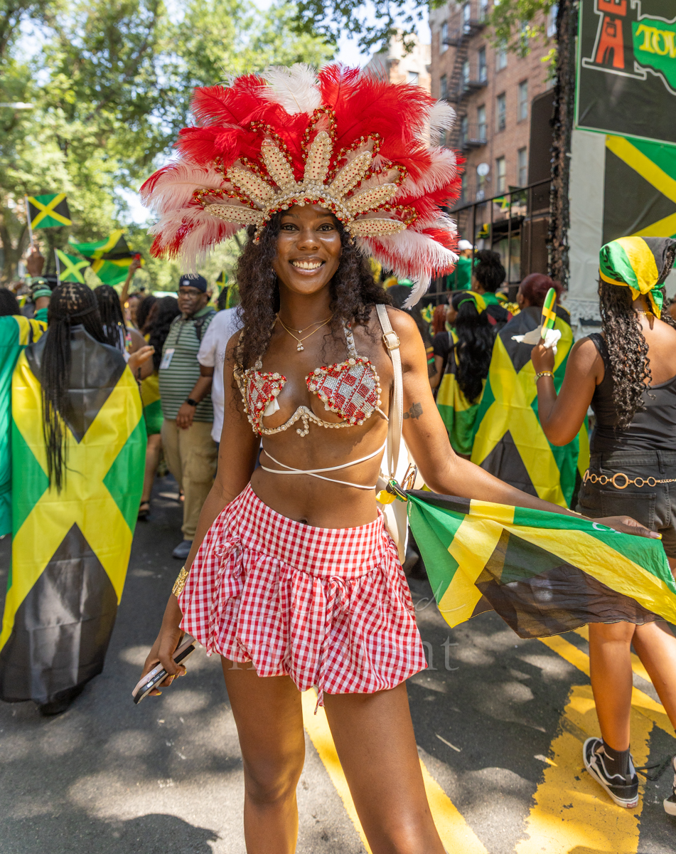 Smiling participant in a red-and-white feathered headdress and beaded carnival costume holds a Jamaican flag