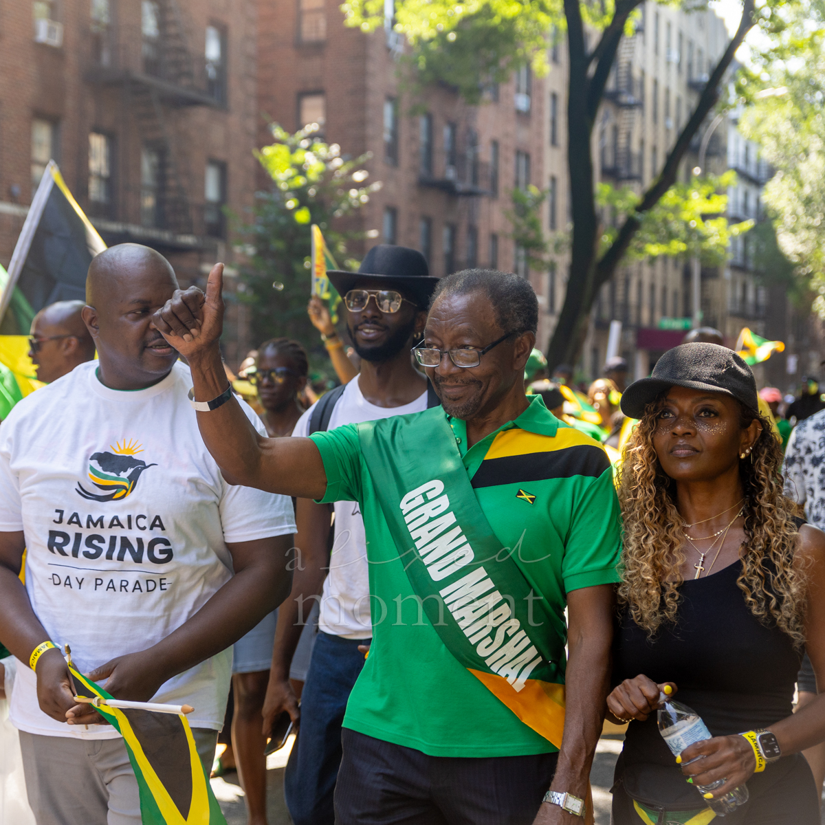 Male grand marshal in a green Jamaica polo shirt raises one hand while walking beside parade participants