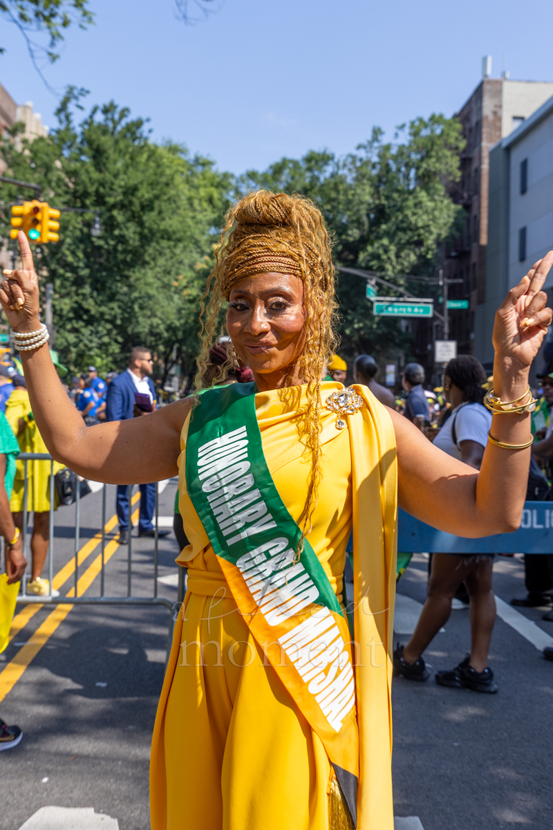 Honorary grand marshal in a gold dress and green sash raises both hands on the Brooklyn parade route