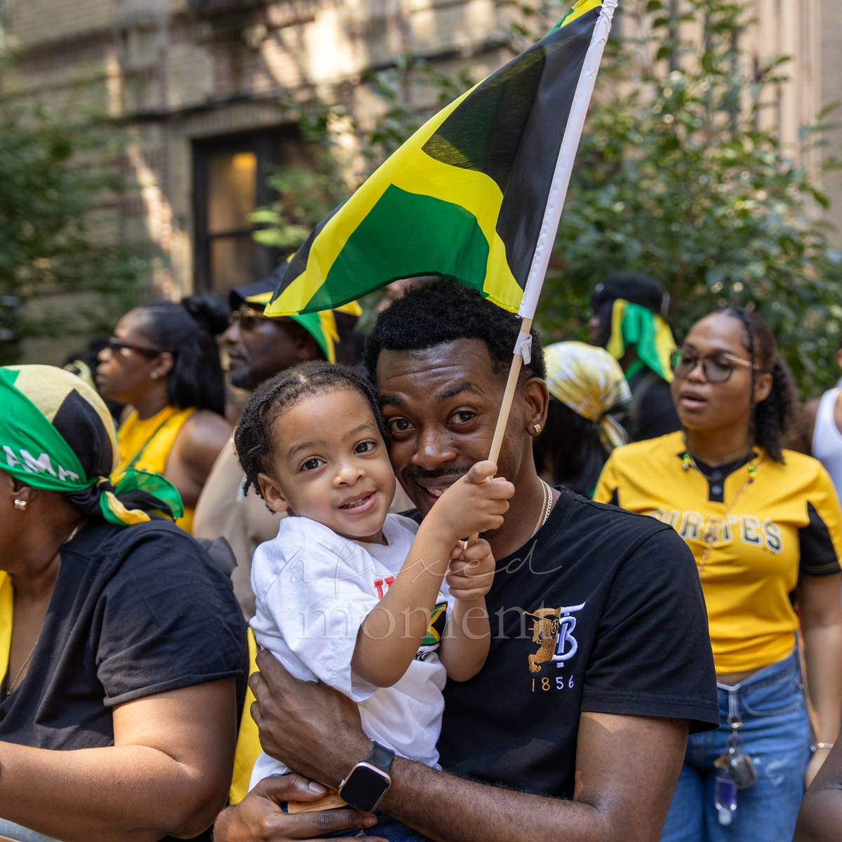 Man carries a young child beneath a large green, gold and black Jamaican flag during the Brooklyn parade