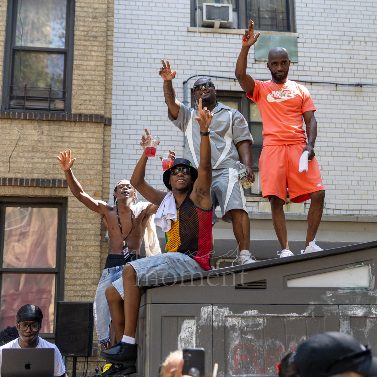 Four men wave and gesture from the roof of a parade sound vehicle while a DJ works below
