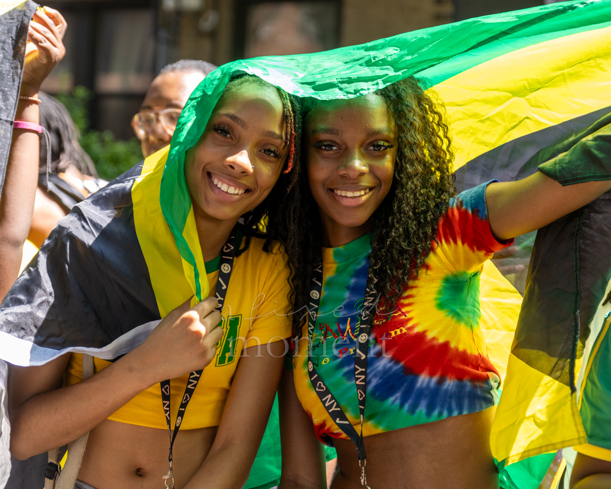 Two smiling young women pose together beneath a large green, gold and black Jamaican flag