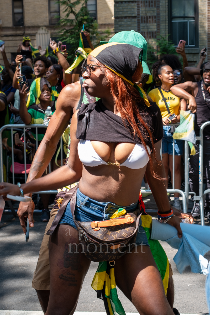 Woman in a white top, denim shorts and Jamaican-colored headscarf dances as spectators record her