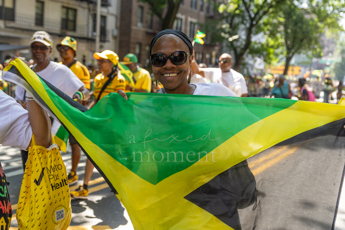 Smiling woman in black sunglasses and a white T-shirt holds a large Jamaican flag open in front of her