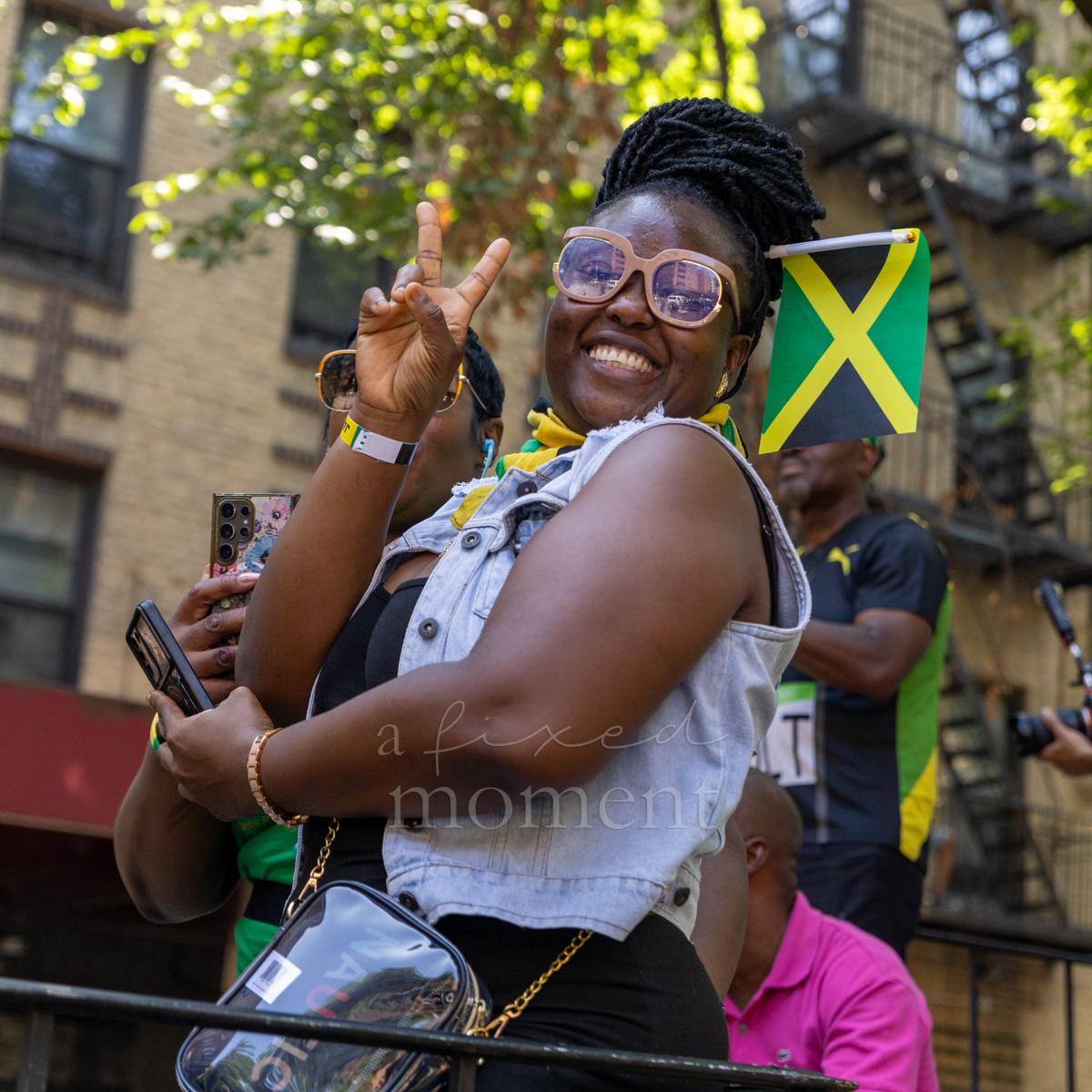 Smiling woman in pink sunglasses and a denim vest flashes a peace sign beside a small Jamaican flag