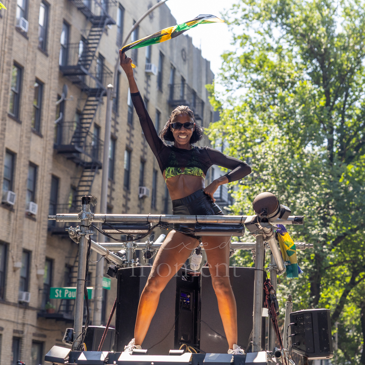 Woman in sunglasses raises a Jamaican flag while standing above speakers and metal rigging on a parade sound vehicle