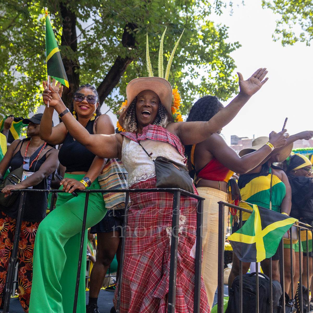 Women celebrate beside Jamaican flags as a participant in a decorated straw hat and red plaid wrap stretches her arms wide