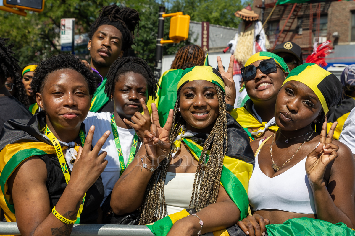 Six young paradegoers draped in Jamaican flags pose together and flash peace signs behind a metal barricade