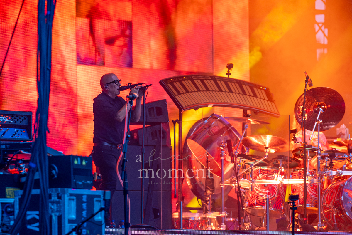 Tool performs against an orange-red backdrop and large drum setup at Riot Fest