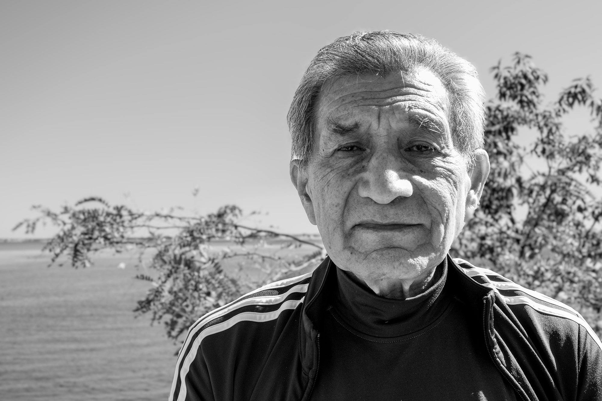 Black-and-white portrait of Pastor Hugo outdoors, with trees and a waterfront in the background.