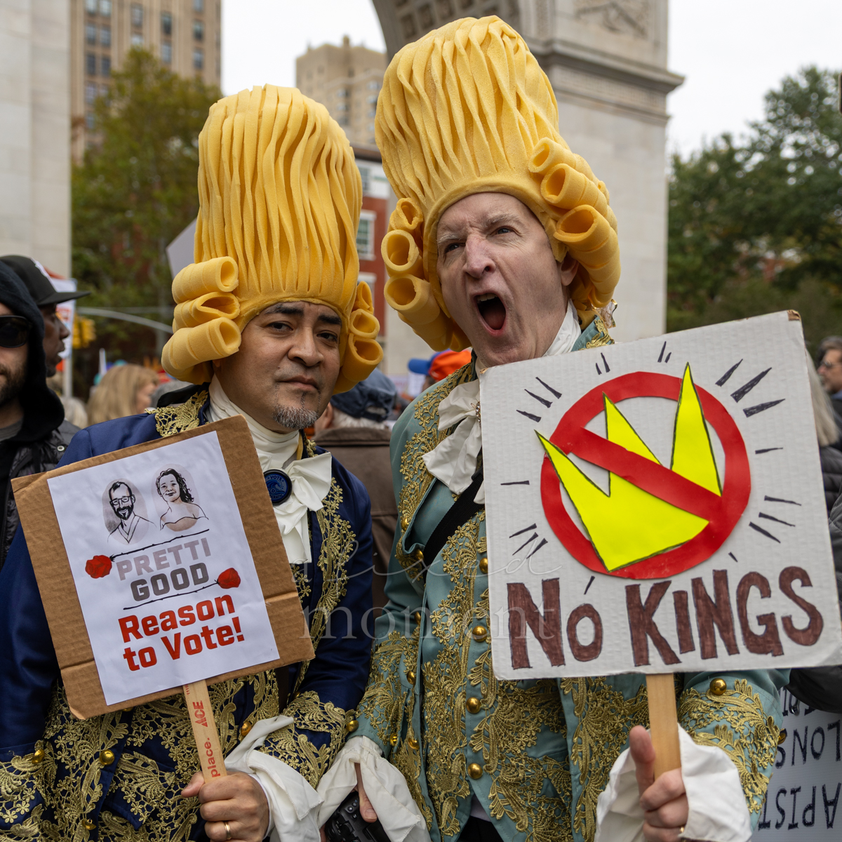 Two attendees wear elaborate yellow wigs and carry No Kings and voting signs.