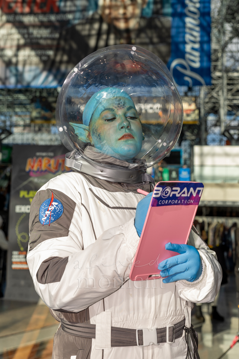 A blue-green alien astronaut cosplayer wears a clear spherical helmet and holds a pink clipboard.