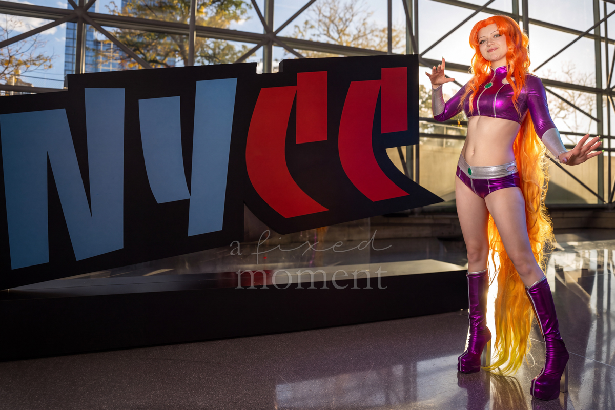 A Starfire cosplayer with long orange hair and a purple costume poses beside the New York Comic Con sign.