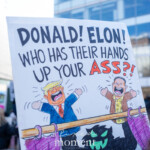 A hand-drawn protest sign is held high during the 50 50 1 march in New York City on February 17, 2025. The sign features caricatures of Donald Trump and Elon Musk depicted as puppets, seemingly controlled by a dark figure below the stage. The text on the sign reads: "DONALD! ELON! WHO HAS THEIR HANDS UP YOUR ASS?!" in bold black and red letters. Trump is illustrated with exaggerated facial expressions, wearing a blue suit with a red tie, while Musk is drawn in a brown jacket and yellow shirt adorned with swastikas. The background of the image features a crowd of protesters, many bundled up in winter clothing, with a cityscape of modern glass buildings behind them. A red flag is visible on the right side of the frame. The protest took place at Union Square and marched towards Washington Square Park, advocating against political and corporate influence.