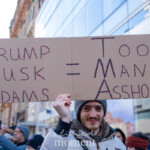 A protester at the 50 50 1 march in New York City on February 17, 2025, holds a large cardboard sign with bold, handwritten text. The sign reads:
"TRUMP MUSK ADAMS = TOO MANY ASSHOLES", with the first letters of each name and phrase highlighted in red to form the acronym "TMA = TMA." The protester, a young man wearing a gray beanie and a light gray winter jacket, smiles while holding the sign high. Behind him, a diverse crowd of demonstrators, many dressed in warm clothing, are gathered. Some participants are taking photos or recording the event with their phones. The background includes modern glass buildings and classic brick architecture, suggesting a location near Union Square, where the march began before heading to Washington Square Park.