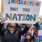 A woman and a young child stand together at the 50 50 1 march in New York City on February 17, 2025, holding a large protest sign that reads: "IMMIGRATION BUILT THIS NATION." The text is handwritten in bold, colorful letters, with "IMMIGRATION" featuring a mix of red, green, blue, and orange, and "NATION" outlined in vibrant shades. The woman wears a black winter coat, a blue scarf, sunglasses, and gloves while carrying a black crossbody bag. She has a bright smile and confidently raises the sign high. The child beside her wears a camo-patterned winter jacket with red, black, and green tones, a shark-toothed beanie, and black gloves. They hold onto the sign as well, looking towards the camera with a determined expression.
In the background, a crowd of demonstrators is visible, many holding signs advocating for various causes. Some participants are bundled up in winter clothing, and a secondary protest sign partially visible behind the main subjects reads: "MAKE AMERICA GREAT" in blue and green letters. The setting appears to be near Union Square, where the march started before heading to Washington Square Park.
