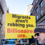 An older protester participates in the 50 50 1 march in New York City on February 17, 2025, holding a bold yellow sign with black and red text. The message on the sign reads: "Migrants aren’t robbing you. Billionaires are." The man is dressed in a gray hoodie, a patterned knit beanie, black gloves, and sunglasses. His expression is serious as he holds the sign high, emphasizing the economic disparity argument being made at the demonstration.
The background captures an urban street scene with classic New York brick and stone buildings lining the street. Other demonstrators, some bundled up in winter clothing, are visible walking and participating in the protest. Vehicles, including trucks and cars, are seen in the distance. The event began at Union Square and continued towards Washington Square Park, drawing attention to social and economic justice issues.