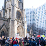 A large group of protesters participates in the 50 50 1 march in New York City on February 17, 2025, demonstrating outside a historic Gothic-style church. The crowd, dressed in winter clothing, holds up various signs and placards, some with messages advocating for social justice and political change. One visible sign reads "SAVE OUR NATIONAL PARKS," while others feature bold colors such as red, green, and white with slogans related to freedom and activism.
The scene takes place in front of a grand stone church with towering spires, arched windows, and intricate architectural details. Behind the church, a modern high-rise residential building contrasts with the historic structure. The protest appears to be in motion, with people raising their signs, engaging in discussions, and moving forward as part of the march, which began at Union Square and headed towards Washington Square Park.