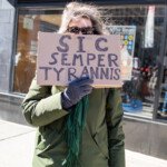 A protester stands on the streets of New York City during the 50 50 1 march on February 17, 2025, holding a cardboard sign with bold, hand-written text that reads: "SIC SEMPER TYRANNIS." The phrase, which translates from Latin to "Thus always to tyrants," is historically associated with resistance against oppressive rule. The protester wears a long olive-green winter coat, a matching green scarf with fringes, dark gloves, and sunglasses. Their windblown hair partially covers their face as they grip the sign firmly.
The background showcases an urban setting with a glass storefront reflecting the bustling street activity. Other demonstrators are visible in the reflection, engaging in the protest. The event, which started at Union Square and continued towards Washington Square Park, focused on various political and social justice issues, drawing a diverse crowd of activists.