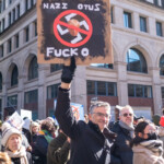 A demonstrator at the 50 50 1 march in New York City on February 17, 2025, raises a bold protest sign featuring a modified Nazi symbol with an image of Donald Trump at its center, crossed out with a red circle. The sign also includes the text "NAZI OTUS" at the top and "FUCK 0" at the bottom, painted in white over a black background. The protester, a middle-aged man with gray hair and glasses, wears a black winter coat and black gloves. He smiles slightly as he lifts the sign high while marching with a crowd of demonstrators.
The background features a dense group of protesters carrying various signs, some of which also display anti-Trump messages. The demonstration takes place in a bustling part of the city, surrounded by historic buildings with large windows. Many attendees are dressed in warm clothing, with some wearing hats, scarves, and gloves, indicative of the chilly February weather. The march, which began at Union Square and headed towards Washington Square Park, is part of a larger movement advocating for political change and social justice.