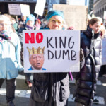 An older protester participates in the 50 50 1 march in New York City on February 17, 2025, holding a sign that reads "NO KING DUMB." The sign features a digitally edited image of Donald Trump wearing a golden crown, emphasizing a critique of authoritarianism. The words "NO" are in red, while "KING DUMB" is in bold black lettering.
The protester wears a teal knitted beanie, yellow-framed glasses, and a dark winter coat with gloves. Behind them, a diverse crowd of demonstrators holds signs with various messages, including one that partially reads "NO ONE VOTED FOR THIS!" The march takes place on a city street lined with high-rise buildings and office spaces, with a mix of pedestrians and activists walking together. The demonstration, which began at Union Square and continued to Washington Square Park, draws attention to concerns about democracy and political power.