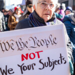 An elderly protester at the 50 50 1 march in New York City on February 17, 2025, firmly holds a sign with a message referencing the U.S. Constitution. The sign reads: "We the People NOT Your Subjects," with "We the People" written in the iconic script from the Constitution and "NOT" emphasized in bold red letters. This message critiques perceived government overreach and emphasizes the principles of democracy and citizen sovereignty.
The protester has white hair and wears a dark winter coat, a multicolored scarf, and transparent-framed glasses. Their facial expression is serious and determined as they stand among a diverse crowd of demonstrators. In the background, other protesters are holding various signs, with some partially visible messages advocating for political and social justice causes. The march, which started at Union Square and proceeded towards Washington Square Park, highlights civic engagement and activism.