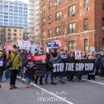 A large group of demonstrators participates in the 50 50 1 march in New York City on February 17, 2025, marching through the streets carrying signs and banners opposing political corruption and authoritarianism. The most prominent banner, held by multiple protesters, reads "STOP THE GOP COUP" in bold white letters on a black background. Smaller signs among the crowd include messages such as "DEFEND THE CONSTITUTION," "NO DICTATORS IN THE USA," and "F'ELON AND THE FELON," referencing Elon Musk and Donald Trump. Many signs feature images of Trump with slogans denouncing his influence and policies.
Protesters wear winter clothing, including hats, scarves, and gloves, as they march along a city street lined with red brick apartment buildings and modern glass structures. One man, wearing a black hat and a mustard-colored coat, raises a sign that reads "DEFEND THE CONSTITUTION." Another protester in a red beanie and black jacket holds a "STOP THE COUP" sign. The crowd is diverse, including people of different ages and backgrounds, all walking together in a peaceful demonstration.
A photographer in the foreground captures the scene, while a security or event marshal in a neon yellow vest observes the march. The event, which began at Union Square and headed toward Washington Square Park, represents a broader movement advocating for democracy and resistance against perceived political threats.