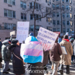 A diverse group of demonstrators participates in the 50 50 1 march in New York City on February 17, 2025, walking through a residential area while holding protest signs and banners. Two prominent signs in the image read: "ISO A MAYOR WHO ISN’T A PUPPET" and "HE WHO SAVES HIS COUNTRY DOES NOT VIOLATE ANY LAW ORANGE." These messages criticize political figures and advocate for accountability.
A protester in the foreground is wrapped in a trans pride flag (light blue, pink, and white), symbolizing LGBTQ+ solidarity within the movement. Another marcher, wearing a black winter coat and hat, gestures as they engage with the crowd. The protesters are bundled in warm clothing, including hats, gloves, and heavy coats, indicating cold weather conditions.
The demonstration takes place on a city street lined with apartment buildings. In the background, more protesters carry various signs with messages advocating for democracy, resistance against authoritarianism, and social justice. The march, which started at Union Square and continued toward Washington Square Park, is part of a larger movement pushing for political change and equity.