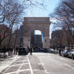 A view of the Washington Square Arch in New York City, marking the endpoint of the 50 50 1 march on February 17, 2025. The iconic marble structure stands tall, framed by leafless trees, as a crowd of demonstrators gathers beneath it. The late winter sun shines through the branches, casting long shadows on the street.
In the foreground, a pedestrian in a long coat walks toward the arch, while parked cars and NYPD vehicles are visible along the curbs. Protesters are seen mingling near the entrance to Washington Square Park, some carrying signs and banners. The presence of police officers and barricades indicates crowd management efforts. This moment captures the conclusion of the march, where participants reflect on the day's activism and advocacy.