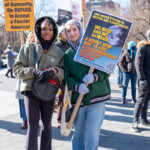 Two young protesters stand together at the 50 50 1 march in New York City on February 17, 2025, holding signs advocating for systemic change and resistance against fascism. One protester, wearing a green hoodie under an oversized khaki jacket, holds a compact black drum with drumsticks tucked inside. The other, wearing a green and white varsity jacket with a gray headscarf, holds a sign that reads: "THIS WHOLE SYSTEM IS ROTTEN AND ILLEGITIMATE! WE NEED AND WE DEMAND A WHOLE NEW WAY TO LIVE A FUNDAMENTALLY DIFFERENT SYSTEM" with a QR code linking to revcom.us (@therevcoms).
Behind them, other demonstrators hold up signs, including a bold yellow one reading: "In the Name of Humanity, We REFUSE to Accept a Fascist America!" from RefuseFascism.org. Another partially visible sign states, "NO DICKTATORSHIP!", adding to the political messages being shared. The event takes place in Washington Square Park, with a diverse group of activists standing together under the bright winter sun, dressed in warm layers, scarves, and gloves.