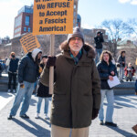 An older protester stands in Washington Square Park during the 50 50 1 march on February 17, 2025, holding a bright orange sign that reads: "In the Name of Humanity, We REFUSE to Accept a Fascist America!" The sign is branded with the RefuseFascism.org website, emphasizing a movement against authoritarianism and political oppression.
The protester wears a thick olive-green winter coat with a fur-lined hood, a gray knit beanie, glasses, and gloves to protect against the cold. His determined expression and upright posture reflect his commitment to the cause. Behind him, more demonstrators are holding similar Refuse Fascism signs, engaging in discussions, and standing in solidarity. The setting is vibrant with people of all ages, some carrying pamphlets or additional protest materials. A woman in a dark blue jacket is seen speaking while holding documents, and another protester in a black hoodie observes the scene.
The protest, which began at Union Square and marched towards Washington Square Park, gathered activists advocating for democracy, resistance against authoritarianism, and social justice.