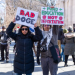 Two demonstrators stand in Washington Square Park during the 50 50 1 march on February 17, 2025, holding up protest signs addressing political and social issues. The woman on the left, dressed in a long black puffer coat, sunglasses, and jeans, holds a white sign with bold red and black text reading: "BAD DOGE." The message appears to be a play on words, potentially referencing political figures or policies.
The protester on the right wears a gray jacket, a keffiyeh scarf, and purple-tinted glasses, holding a sign that reads: "FUND EDUCATION NOT DEPORTATION" in green and red, with symbols opposing deportation policies. A black crossbody bag with a red design hangs from their shoulder, and a black instant camera is strapped around their neck. They also wear gloves and brown leather shoes, indicating cold weather conditions.
The background features a crowd of demonstrators, some holding additional protest signs, with leafless trees and historic statues in Washington Square Park. The march, which started at Union Square, highlights issues such as immigration, education funding, and government accountability.
