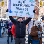 A demonstrator at the 50 50 1 march in New York City on February 17, 2025, raises a large handmade sign that reads "SAVE OUR NATIONAL PARKS!" The sign features hand-drawn images of trees, a giant sequoia, and a park ranger hat with the message "Only WE can SAVE them!!!" in red and green lettering, emphasizing a call to protect the environment and public lands.
The protester wears a black jacket, gray beanie, and black gloves, lifting the sign high above their head with a determined expression. Surrounding them are fellow demonstrators, bundled up in winter clothing, including scarves, coats, and gloves. To the right, a woman in a brown fuzzy coat, sunglasses, and gray gloves walks forward, carrying a navy tote bag. Behind them, other activists hold various protest signs advocating for different causes, while some take photos or engage in conversations. The march, which started at Union Square and ended at Washington Square Park, highlights political activism, environmental advocacy, and public engagement.