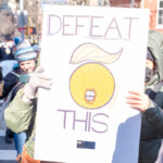 Two demonstrators at the 50 50 1 march in New York City on February 17, 2025, hold up a handmade protest sign featuring the words "DEFEAT THIS" in large, bold letters. Below the text, an illustration of an orange with Trump-like hair and a wide-open mouth symbolizes opposition to the former president. The sign also includes the hashtag #WeWorkHere written on the side.
The protesters, bundled up in winter clothing, wear gloves, jackets, and scarves to shield themselves from the cold. The demonstrator on the left, wearing a green coat and gray gloves, holds the sign with both hands, while the person on the right, dressed in a dark hooded coat and a face mask, supports the other side. The background features a bustling protest scene with more demonstrators holding up signs and banners as they march through the streets of Manhattan. Leafless trees, red-brick buildings, and streetlights frame the setting, indicating a wintertime demonstration.
The 50 50 1 march, which began at Union Square and proceeded to Washington Square Park, is a demonstration advocating for social justice, democracy, and political accountability.