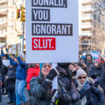 A protester at the 50 50 1 march in New York City on February 17, 2025, firmly holds a large sign with the bold message: "DONALD, YOU IGNORANT SLUT." The sign is designed with a modern, high-contrast layout—black and white text with the word "SLUT." emphasized in a red box, reminiscent of meme and media-style protest art. This phrase is a reference to a well-known Saturday Night Live (SNL) sketch, repurposed here as a political statement against former president Donald Trump.
The protester wears a long camouflage-patterned winter coat, a bright teal beanie, and black gloves, gripping the sign with both hands while looking directly at the camera with a determined expression. The surrounding crowd is filled with demonstrators in winter clothing, many holding signs, engaging in discussions, and marching together. In the background, protesters carry additional signs, including one with a transgender pride flag, further highlighting the diversity of causes represented at the march. The setting, a city street lined with tall buildings and traffic lights, underscores the urban landscape of the demonstration, which started at Union Square and proceeded to Washington Square Park.