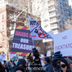 A group of demonstrators at the 50 50 1 march in New York City on February 17, 2025, raises a variety of protest signs and banners criticizing former President Donald Trump and his political allies. Among the most prominent signs:
"NO DICKTATOR", written in bold red text on a white sign, a play on words condemning authoritarian leadership.
"HANDS OFF OUR TREASURY", written in white and red on a black background designed to look like caution tape, highlighting concerns over financial policies and government spending.
A small flag that reads "NOT MY PRESIDENT 45", with additional words such as "liar", "insurrectionist", and "criminal", featuring a red crossed-out circle over the number 45.
Protesters are seen wearing black gloves, winter coats, and hats, with some wearing sunglasses to shield their eyes from the cold winter sun. The background showcases a dense urban environment with tall residential buildings, traffic lights, and leafless trees, emphasizing the setting in Manhattan. The march, which began at Union Square and concluded at Washington Square Park, reflects a powerful stance against authoritarianism, economic injustice, and political corruption.