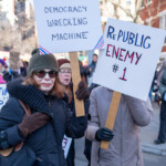Two demonstrators at the 50 50 1 march in New York City on February 17, 2025, hold up handmade protest signs conveying political messages. The sign on the left reads: "DOGE IS A DEMOCRACY WRECKING MACHINE," referring to Elon Musk and the influence of his platforms on political discourse. The sign has decorative red and blue corner accents, giving it a patriotic theme. The protester holding this sign is wearing a green knit beanie, sunglasses, a black winter coat, and brown gloves, with a serious expression.
The sign on the right states: "REPUBLIC ENEMY #1," a play on words suggesting a major political or corporate figure as a threat to democracy. The protester holding this sign is dressed in a beige winter coat with black gloves, keeping their face mostly obscured.
The background captures a large crowd of demonstrators, many holding their own signs, with red-brick buildings, leafless trees, and a city street framing the protest scene. The 50 50 1 march, which started at Union Square and proceeded to Washington Square Park, was a mass demonstration advocating for democracy, political accountability, and corporate regulation.