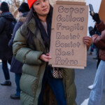 A demonstrator at the 50 50 1 march in New York City on February 17, 2025, holds a handmade cardboard sign with a satirical message that reads: "Super Callous Fragile Racist Sexist Nazi Potus", a play on words referencing Supercalifragilisticexpialidocious from Mary Poppins while critiquing Donald Trump.
The protester is dressed in a long olive-green puffer coat, blue jeans, and a bright orange Carhartt beanie. She also wears gold hoop earrings and carries a black and white checkered purse. Her expression is calm yet assertive as she holds up her sign, making a strong statement against racism, sexism, and authoritarianism.
The background features other protesters in winter attire, some holding their own signs, as they march through the city streets lined with leafless trees and residential buildings. This image captures the essence of the 50 50 1 march, which started at Union Square and proceeded to Washington Square Park, as a movement advocating for democracy, social justice, and political accountability.