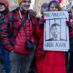 Two protesters at the 50 50 1 march in New York City on February 17, 2025, pose together, making a bold political statement. The woman on the right holds a hand-drawn sign that reads "MAKE AMERICA GREAT AGAIN", featuring an illustration of Donald Trump behind jail bars, a satirical reimagining of his famous slogan.
She is dressed in a bright red puffer coat, matching gloves, and a blue hat, carrying a red crossbody bag. The man beside her, smiling at the camera, wears a red and black plaid jacket, a yellow beanie with black bear ears, and a rainbow lanyard around his neck. He also carries a black Polaroid camera bag across his chest.
The background is filled with other demonstrators holding signs and flags, marching through a city street lined with tall buildings and leafless trees, indicating the winter season. This image captures the spirit of civic engagement and political resistance, with protesters using creative signage and attire to express their views.