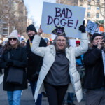 A lively protester at the 50 50 1 march in New York City on February 17, 2025, raises a handmade sign that reads "BAD DOGE!", featuring an illustrated ferocious-looking black dog with sharp teeth. She is energetically marching, smiling broadly, and pointing at the sign with both hands. She is dressed in a white puffer coat, a gray hoodie, black gloves, and black pants, embracing the winter chill.
Surrounding her are fellow demonstrators also participating in the march. To her left, a man in a black beanie, glasses, and a dark coat walks alongside a woman wearing a black coat, beanie, and sunglasses, carrying a black bag. Behind them, a crowd of protesters holds various signs, some of which include political statements and symbolic illustrations. The backdrop consists of a city street lined with tall buildings and leafless trees, a testament to the winter season.
This image captures the energy, unity, and defiance of the protesters, as they voice their stance against political figures and policies they oppose.