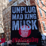 A demonstrator at the 50 50 1 march in New York City on February 17, 2025, holds a bold black protest sign that reads "UNPLUG MAD KING MUSK" in large, distressed white text. Below the statement is a red circle containing a black-and-white image of Elon Musk's face, and the hashtag #NoKingsDay is displayed prominently at the bottom in red letters.
The surrounding crowd is filled with protesters raising various signs, including one that features a red sign with a question mark and the letters "WTF?" The protest is taking place in a city street lined with red brick buildings and leafless trees, indicating the winter season. The event reflects a strong anti-oligarchy and pro-democracy sentiment, with demonstrators calling for the end of excessive power and influence by billionaires.
This powerful image captures the political activism and resistance against corporate control, as demonstrators challenge figures they believe hold too much economic and social power.
