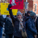 A woman participating in the 50 50 1 march in New York City on February 17, 2025, raises a red protest sign with the bold text "WTF?" alongside an image of Elon Musk smirking and waving. The sign includes handwritten text below Musk’s image, appearing to criticize his policies or actions. She is smiling while wearing a dark winter coat, a colorful polka-dot scarf, a gray knit hat, and black gloves.
In the background, other demonstrators hold signs, including one yellow sign with text referencing immigrants, STEM, and money. The protest takes place on a city street lined with red brick buildings, a one-way street sign, and a security camera mounted on a pole. Protesters are dressed in warm layers, reflecting the winter weather in New York City. The demonstration is part of a broader movement against wealth concentration, political corruption, and billionaire influence in democracy.