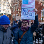 A woman participating in the 50 50 1 march in New York City on February 17, 2025, raises a handmade protest sign featuring an American flag with the words "Real Patriots RESPECT the CONSTITUTION" written in bold red, blue, and black letters. She is wearing a dark blue quilted jacket, a purple knit hat, a matching scarf, and glasses, smiling confidently while holding the sign high.
Beside her, a man in a blue beanie and gray winter coat walks alongside the crowd. Other demonstrators in warm winter clothing are visible in the background, many holding signs. The march takes place in a residential area lined with red brick buildings and leafless trees, typical of winter in New York City. The protest emphasizes democracy, constitutional rights, and political accountability.
