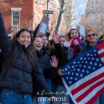 A lively group of young protesters waves and smiles at the camera during the 50 50 1 march in New York City on February 17, 2025. The demonstrators, mostly young women, are dressed in black puffer jackets and jeans, with one person on the left wearing a white knit beanie. A young man in the background, wearing a mustard-colored beanie and sunglasses, holds up an American flag as the group marches together. Behind them, other protesters can be seen holding signs as they move through the city streets lined with red brick buildings and bare trees.
The atmosphere is energetic and optimistic, with participants advocating for democracy and civic engagement. This moment captures a sense of unity and determination among the younger generation, emphasizing their active role in social movements.