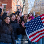A lively group of young protesters waves and smiles at the camera during the 50 50 1 march in New York City on February 17, 2025. The demonstrators, mostly young women, are dressed in black puffer jackets and jeans, with one person on the left wearing a white knit beanie. A young man in the background, wearing a mustard-colored beanie and sunglasses, holds up an American flag as the group marches together. Behind them, other protesters can be seen holding signs as they move through the city streets lined with red brick buildings and bare trees.
The atmosphere is energetic and optimistic, with participants advocating for democracy and civic engagement. This moment captures a sense of unity and determination among the younger generation, emphasizing their active role in social movements.